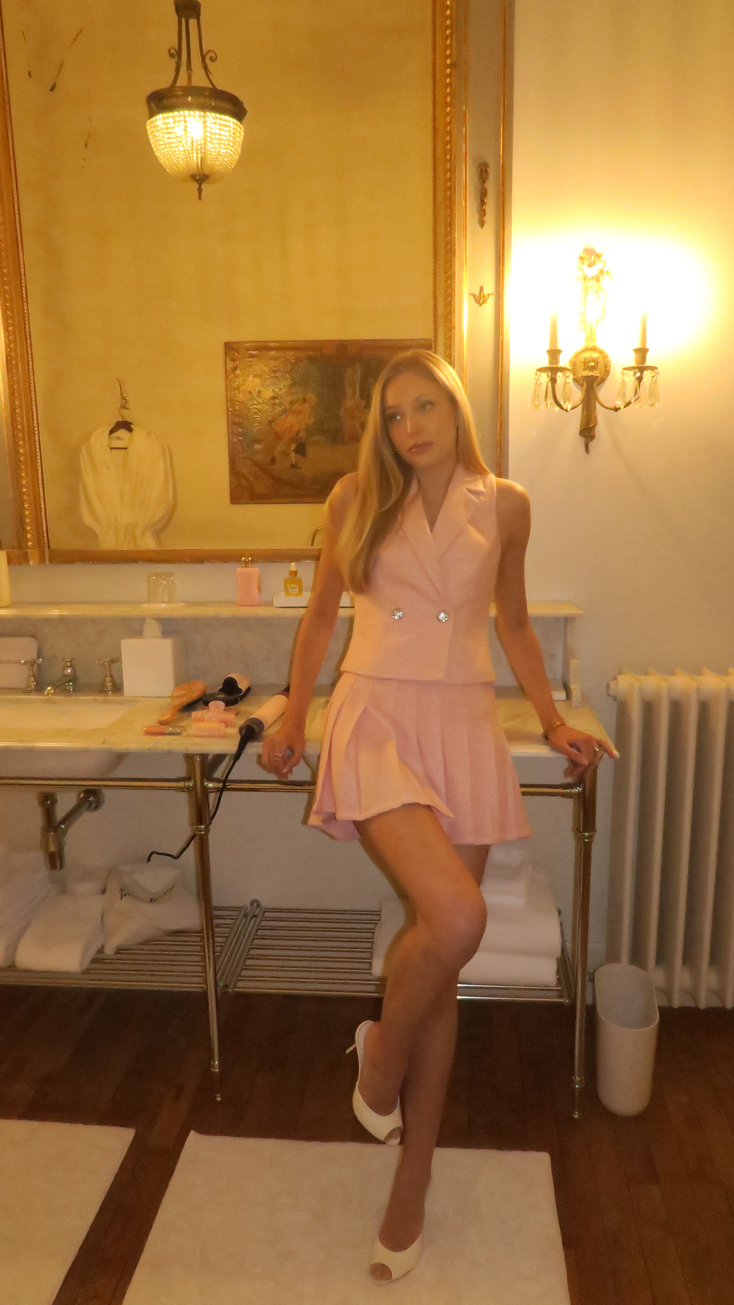 A person wearing a baby pink high waisted, pleated mini skirt sitting on a table in a room with gold-colored walls and a painting in the background.
